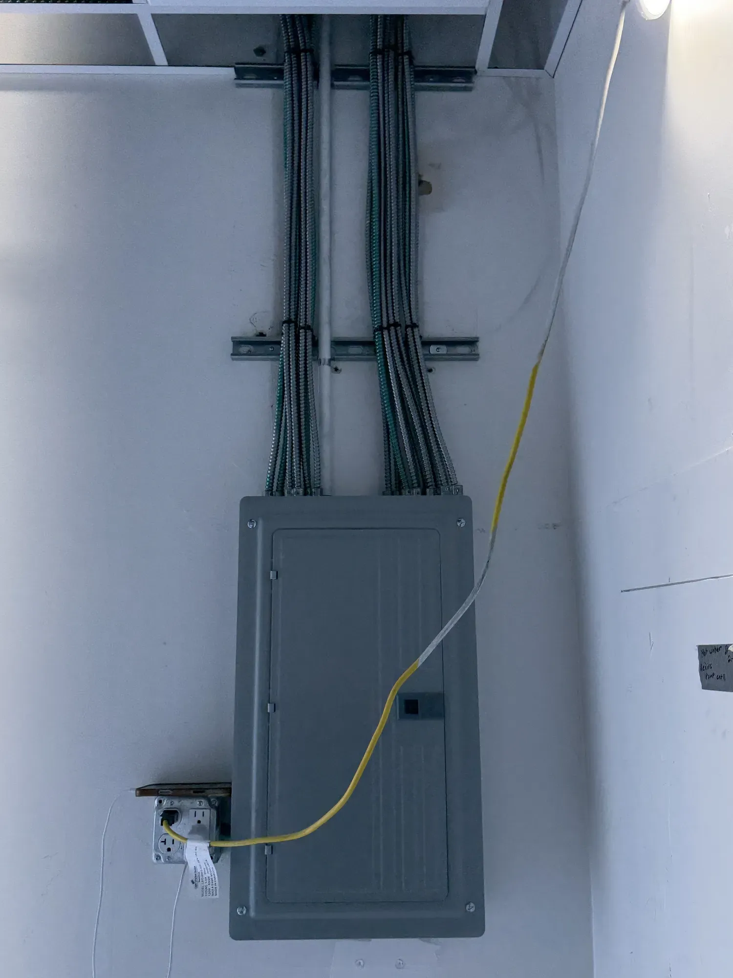 A gray breaker panel mounted on a garage wall with several conduits running up from it to the ceiling