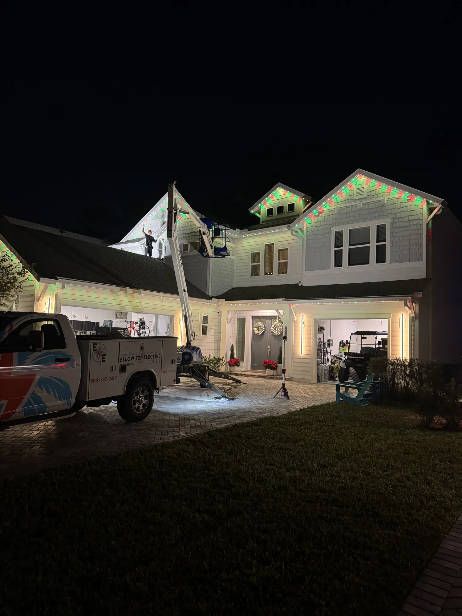 Permanent roofline lights glowing red and green on a two-story home at night while a lift raises the installer to the gable