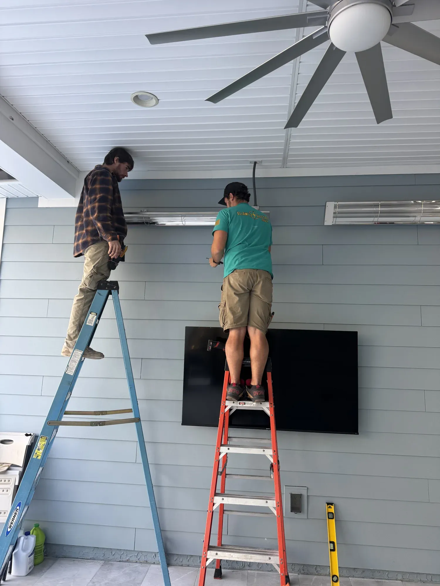 Two Ellowitz electricians on ladders installing infrared patio heaters and a TV at a commercial clubhouse